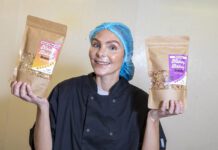 Nakey Bakey secures Scotmid listings Megan Murrie, founder of Nakey Bakey, stands holding two of the brand's granola packs with a hair net and overalls on.