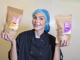 Nakey Bakey secures Scotmid listings Megan Murrie, founder of Nakey Bakey, stands holding two of the brand's granola packs with a hair net and overalls on.
