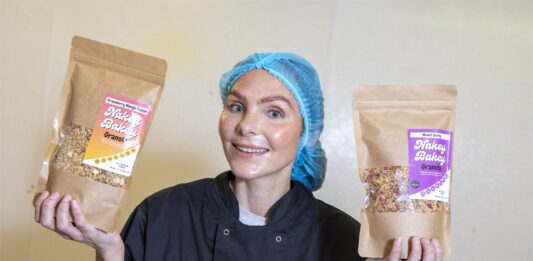 Nakey Bakey secures Scotmid listings Megan Murrie, founder of Nakey Bakey, stands holding two of the brand's granola packs with a hair net and overalls on.