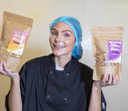 Nakey Bakey secures Scotmid listings Megan Murrie, founder of Nakey Bakey, stands holding two of the brand's granola packs with a hair net and overalls on.