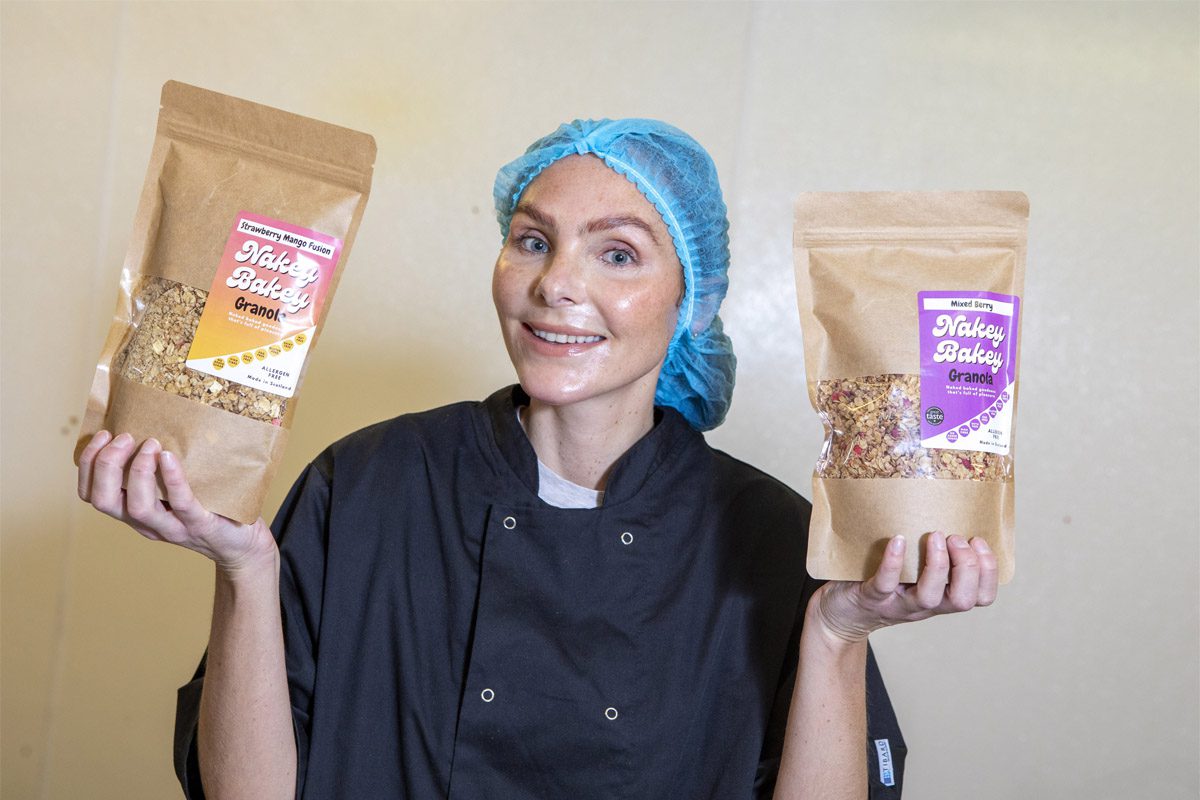Megan Murrie, founder of Nakey Bakey, stands holding two of the brand's granola packs with a hair net and overalls on.