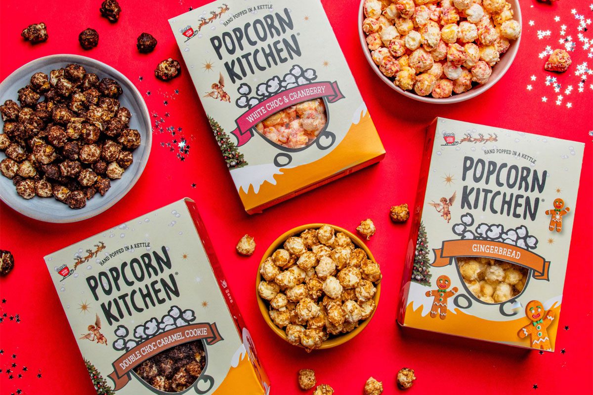 Packs of Popcorn Kitchen Christmas gift packs lie across a red tabletop with bowls of the popcorn around them. The flavorus include White Choc & Raspberry, Double Choc Caramel Cookie and Gingerbread.