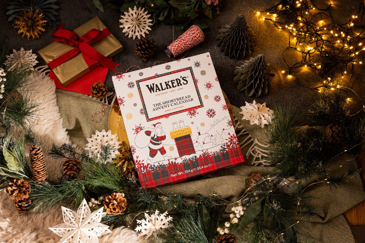 A Walker's Shortbread Advent Calendar sits in the middle of a Christmas scene with presents, decorations and lights.