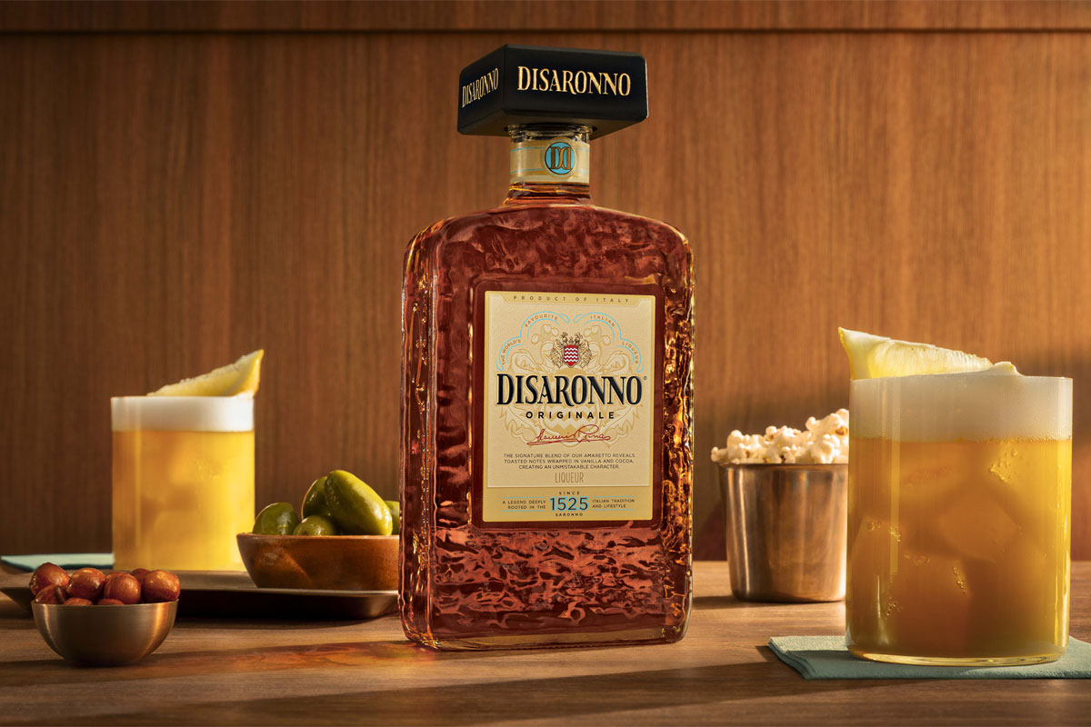 A bottle of the 2026 redesign for Disaronno stands on a table with Amaretto Sour cocktail serves around it as well as bowls of nibbles.