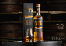 Glasgow 1770 unveils first 10 Year Old peated malt A bottle of Glasgow 1770 10 Year Old Peated Single Malt Scotch Whisky stands on a barrel with its outer packaging next to it with a nosing glass with the whisky inside.