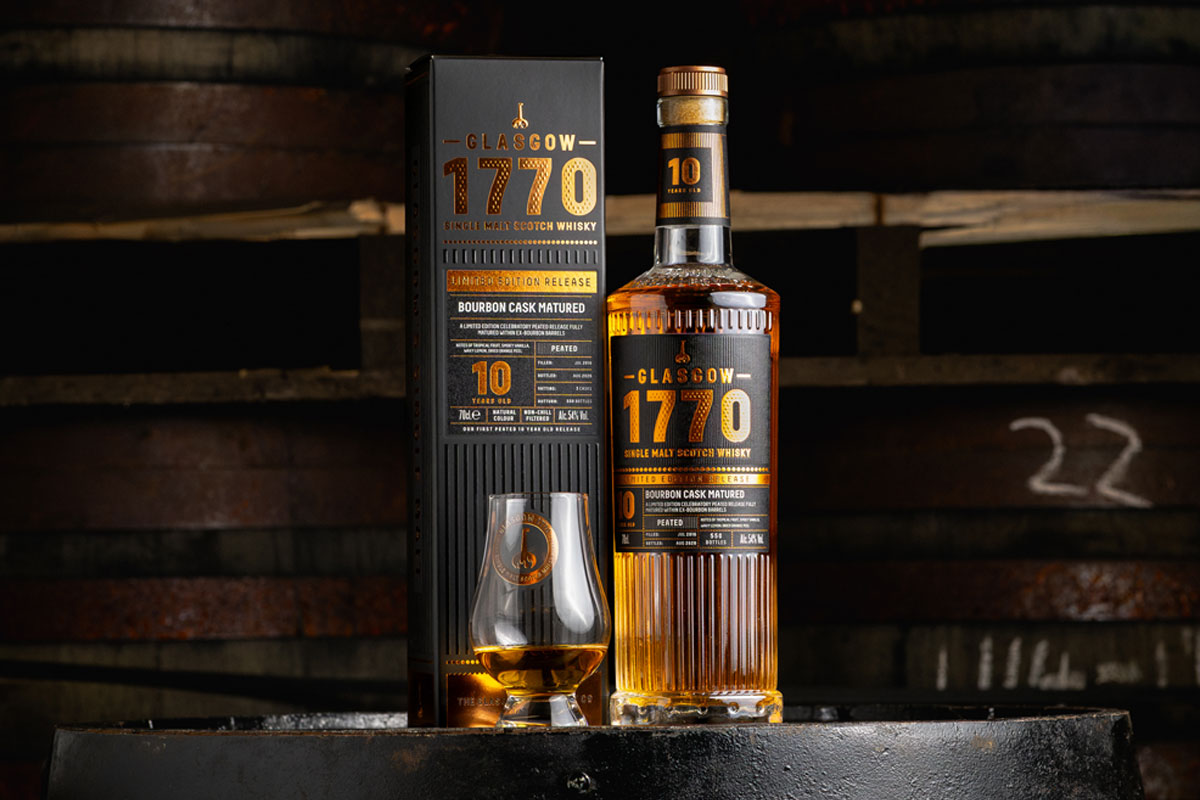 A bottle of Glasgow 1770 10 Year Old Peated Single Malt Scotch Whisky stands on a barrel with its outer packaging next to it with a nosing glass with the whisky inside.