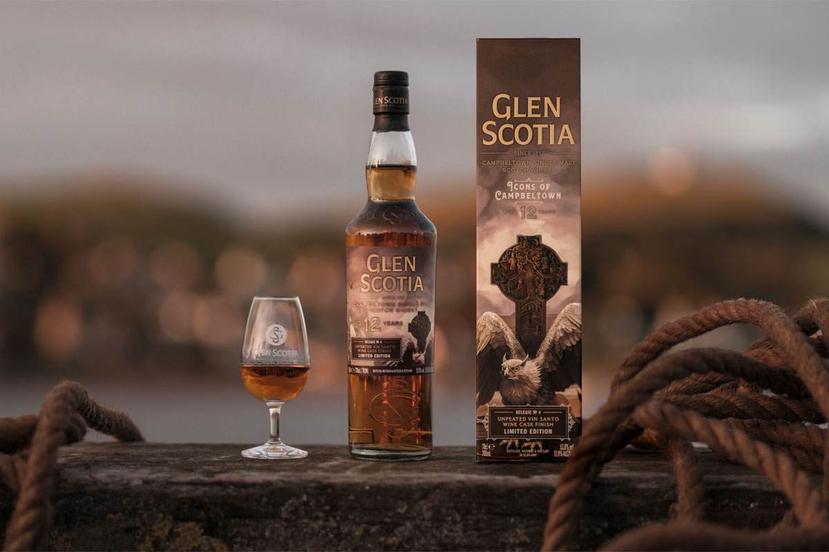 A bottle of Glen Scotia Icons of Campbeltown No.4 stands on a barrel on a dock with the outer case next to it and a nosing glass on its other side with the whisky inside.