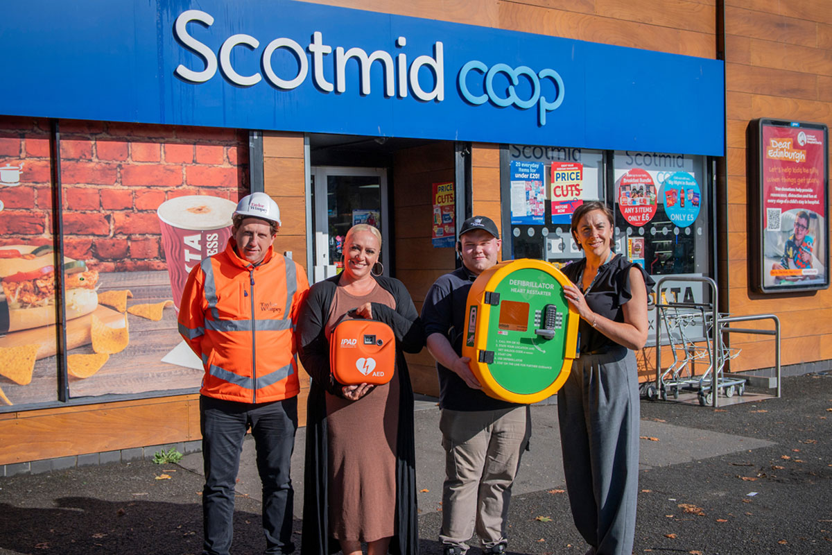 A group of people stand outside Scotmid Ratho Station holding a defibrillator device that is to be installed on the store's exterior wall.