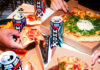 Find a ‘WKD Side’ for Freshers and Halloween A group of people are taking slices of pizza from different boxes with cans of WKDX around the boxes.