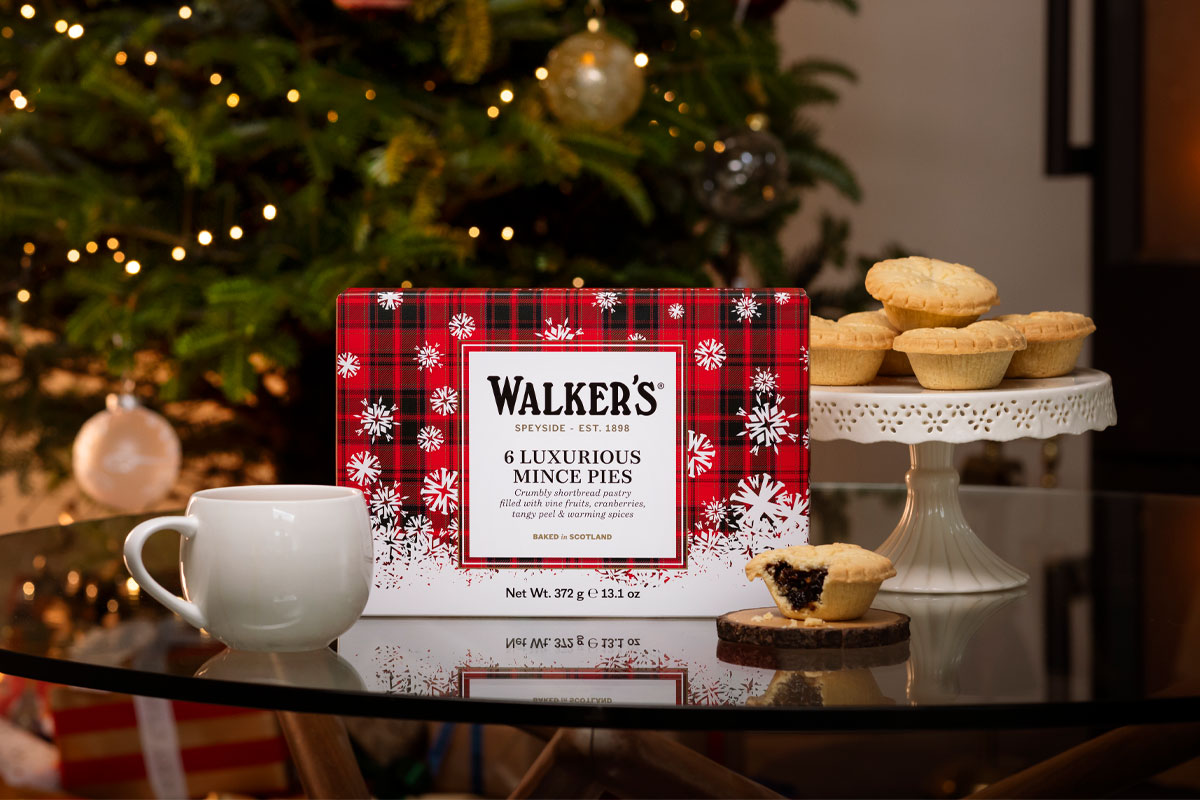 A pack of Walker's Luxury Mince Pies sits on a table with a mug next to it and one of the mince pies to the other side of it. In the background, the pies are stacked up on a table and there is a Christmas tree.