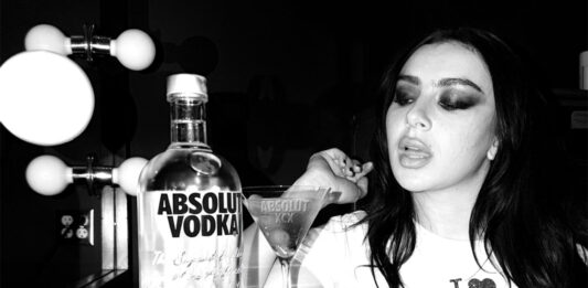 Absolut Vodka teams up with Charli XCX Pop star Charli XCX sits at a dressing table with a Dirty Martini cocktail serve next to a bottle of Absolut Vodka. The photo is in black and white.