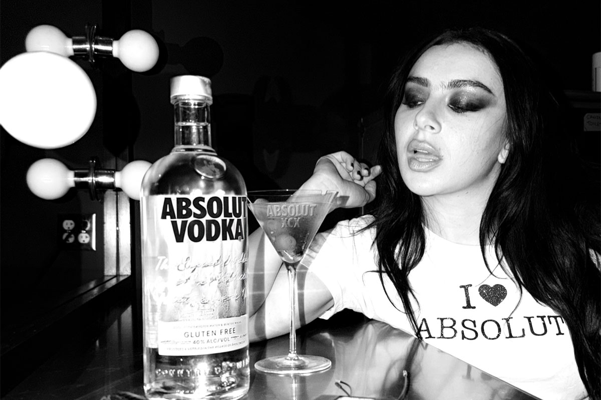 Pop star Charli XCX sits at a dressing table with a Dirty Martini cocktail serve next to a bottle of Absolut Vodka. The photo is in black and white.
