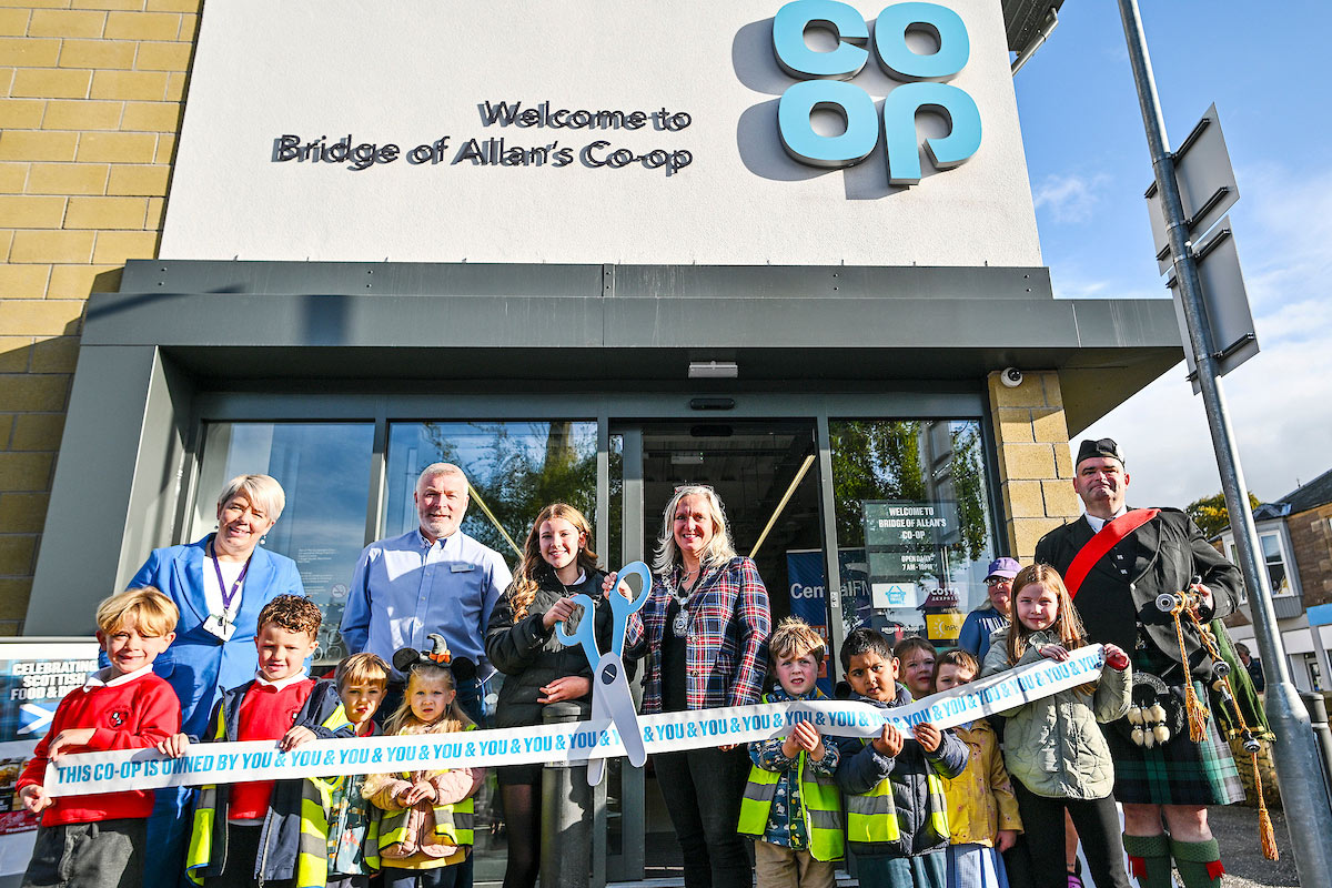 A group of people stand outside Co-op Bridge of Allan holding novelty scissors and cutting a Co-op branded ribbon as part of the store's opening ceremony.