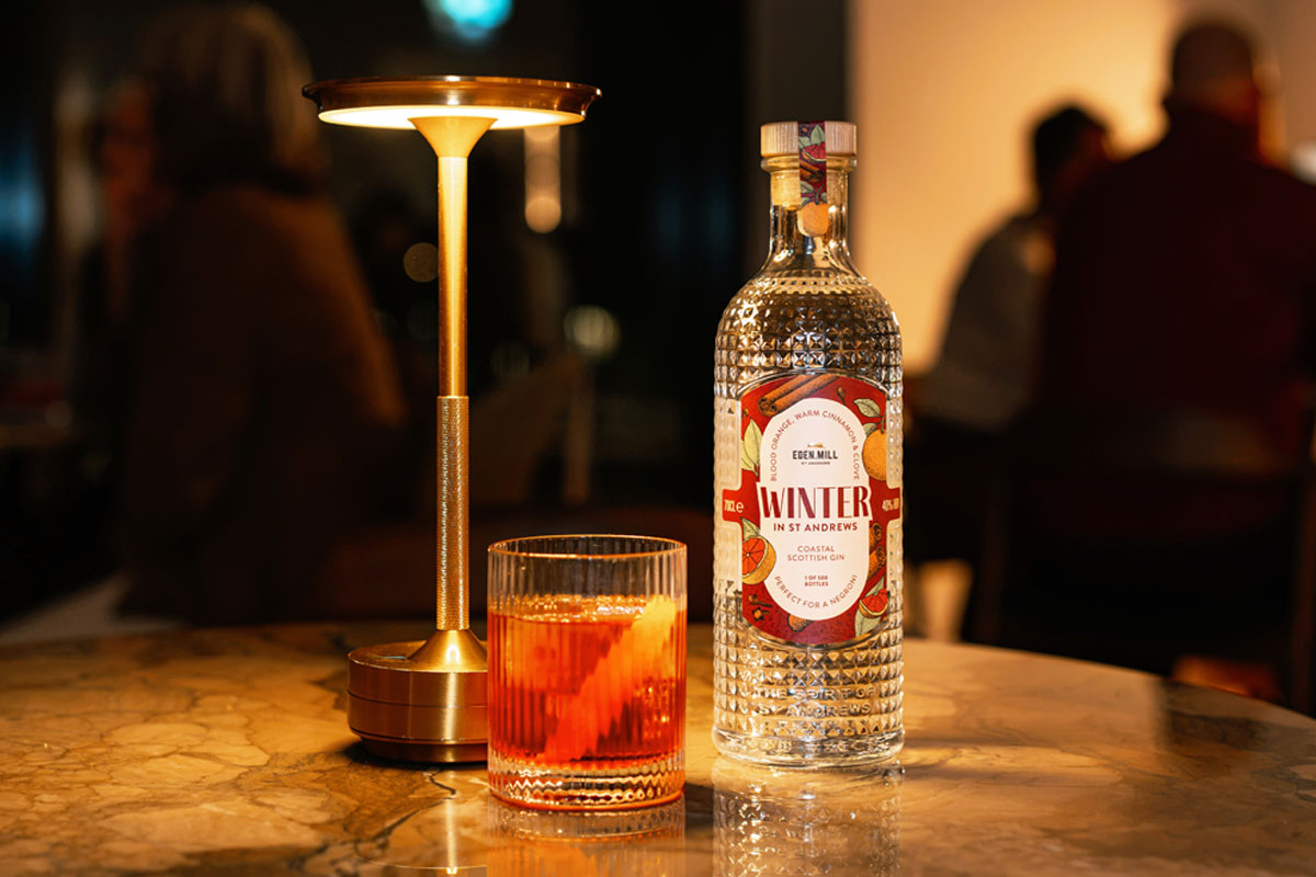 A bottle of Eden Mill Winter in St Andrews gin stands on a table with a Negroni cocktail serve next to it and a lamp.