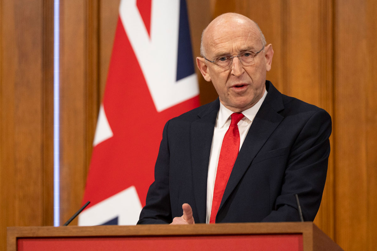 John Healey, Chancellor of the Exchequer, stands at a podium delivering a speech with the UK flag behind him.