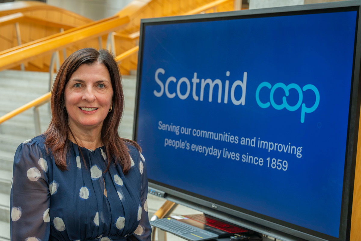Karen Scott, chief executive at Scotmid, stands in Scottish Parliament next to a TV screen with the Scotmid Co-op logo on it.