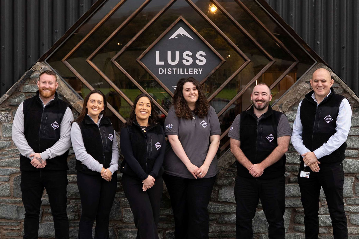 Team members from the Luss Distillery stand outside the building with the Luss Distillery logo behind them.