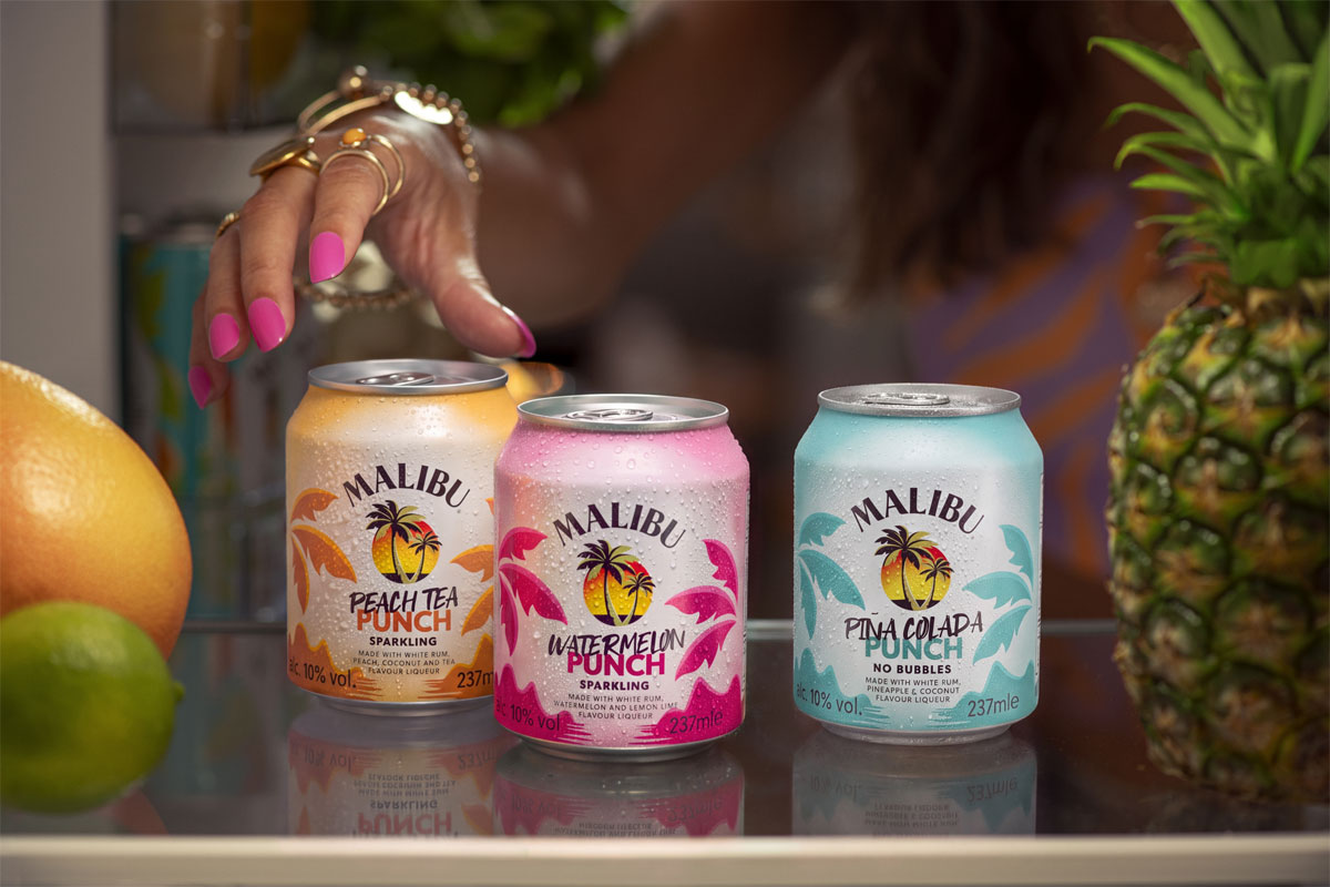 Cans of Malibu Punch sit on a bar as a person's hand reaches for them. There is also different pieces of fruit across the bar.