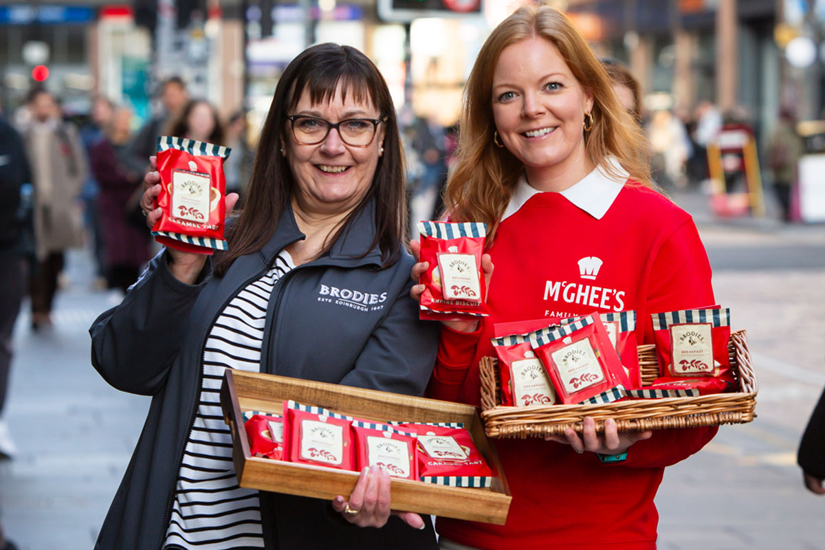 Mina MacKenzie, retail partner manager at Brodies, and Aisling McGhee, commercial director at McGhee's, stand outside Glasgow Central Station holding packs of McGhee's individual biscuits with Brodies tea bag attached to it. Each also hold a wicker basket with more biscuit and tea combos.