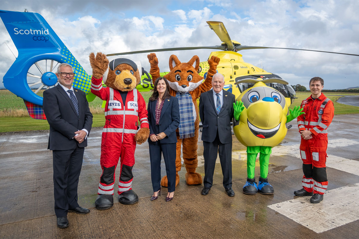 Scotmid Co-op representatives and members of Scotland's Charity Air Ambulance stand on a heliport with their respective mascots and a helicopter behind them.