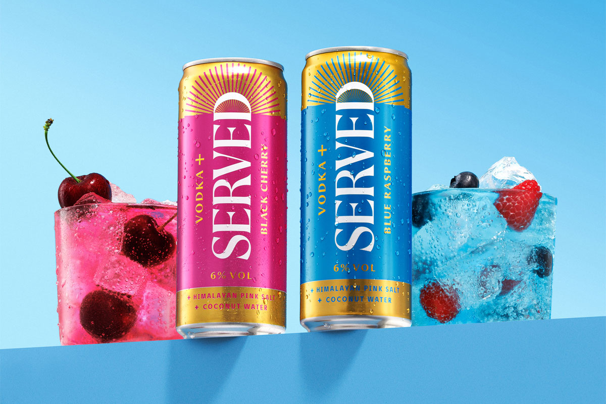 Cans of Served Vodka+ stand on a blue table against a blue background with the drinks poured out into glasses next to them with pieces of fruit inside.