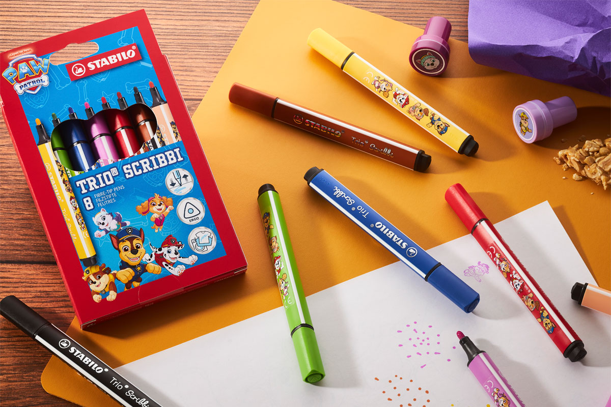 A pack of Stabilo Paw Patrol Trio Scribbi pens sits on a desk with the pans scattered across it with other stationary and pieces of paper.