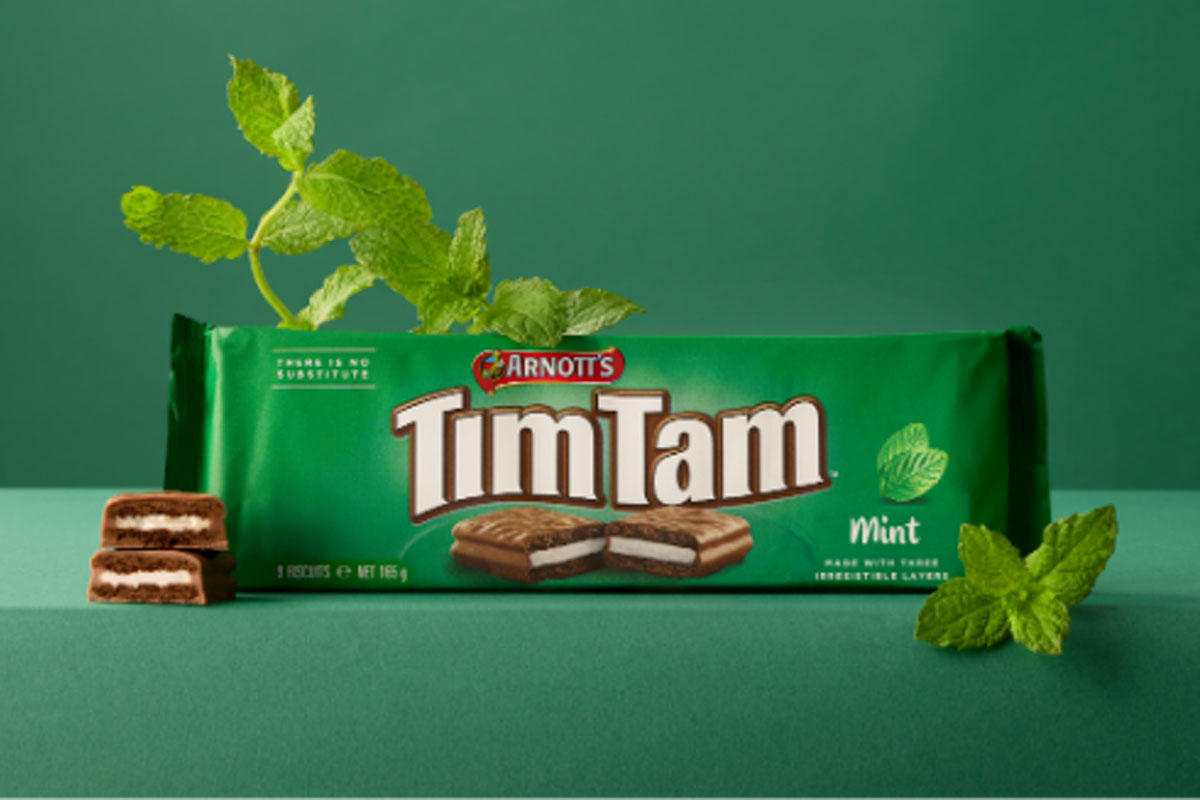 A pack of Arnott's Tim Tam Mint sits on a green table against a green background with mint sprigs around the pack.